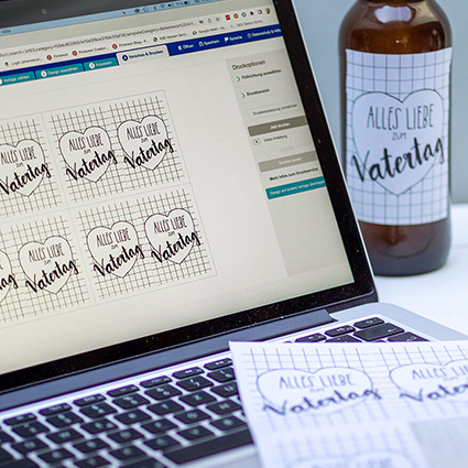 Ein Laptop steht aufgeklappt auf einem Tisch. Auf dem Bildschirm ist die Freeware Design & Print von Avery Zweckform geöffnet. Eine Vorlage für selbstgestaltete Bieretiketten für den Vatertag ist zu sehen. Auf dem Computer liegt ein ausgedruckter Bogen Etiketten. Im Hintergrund steht eine Bierflasche mit eben diesem Etikett. Foto: Johanna Rundel