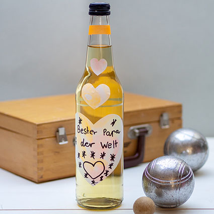 Vor einer hellblauen Wand auf einem hellen Untergrund liegt ein Boule-Koffer aus Holz. Davor steht eine Limonadenflaschen mit einem selbstgestalteten Etikett von Avery Zweckform in Herzform. Darauf ist ein Spruch zum Vatertag geschrieben. Ein paar Boule-Kugeln liegen neben der Flasche. Foto: Johanna Rundel