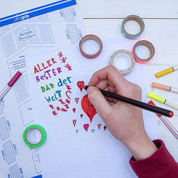 Eine Kinderhand malt ein rotes Herz auf ein Etikett von Avery Zweckform. Drumherum sind bunte Filzstifte und Tape-Rollen verteilt. Foto: Johanna Rundel