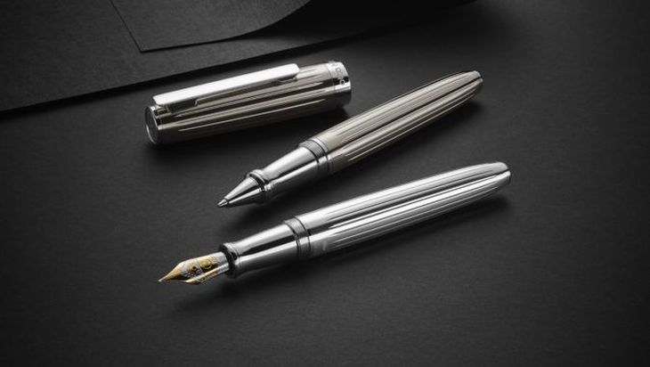 Auf einem schwarzen Hintergrund liegen zwei silberfarbene Stifte: Ein Kugelschreiber und ein Tintenroller. Beide Schreibgeräte sind geöffnet und eine Stiftkappe liegt daneben.
