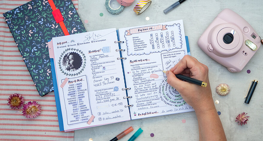 Auf einem hellgrauen Untergrund liegt ein aufgeschlagenes Clipbook von Filofax. Jemand füllt es mit einem dunklen Kugelschreiber aus. Um das Buch herum liegen verschiedene Gegenstände verteilt, die beim Ausfüllen hilfreich sind: eine hellrosa Sofortbildkamera, verschiedene Filzstifte, Washitapes und ein Notizbuch. Foto: Johanna Rundel
