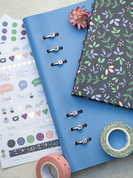 Auf einem hellgrauen Untergrund liegt ein hellblaues Clipbook von Filofax. Es handelt sich um ein Freundebuch für Erwachsene. An den einzelnen Bindungsclips sind kleine Perlen aufgefädelt, die zusammen das Wort "Friend" ergeben. Ein dunkles Notizbuch mit Blumendekor liegt darauf. Daneben ein paar passende Aufkleberbögen und verschiedene Washitapes. Foto: Johanna Rundel