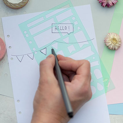 Jemand malt mit einem schwarzen Fineliner und einer Schablone eine Girlande auf eine weiße Filofax-Seite. Darunter liegen weitere Seiten in verschiedenen Pastellfarben. Drumherum sind Blüten und Washitaperollen verteilt. Foto: Johanna Rundel