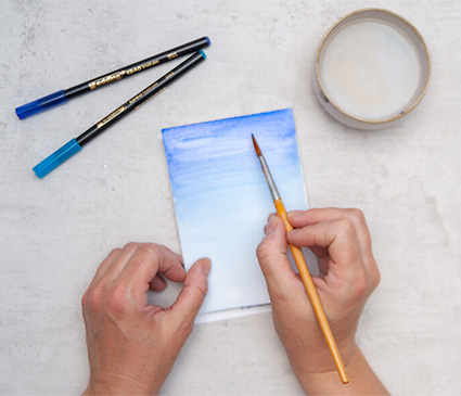 Jemand malt mit einem Pinsel einen blauen Untergrund mit Verlauf von dunkel (oben) nach hell (unten) auf eine Postkarte. Links liegen zwei schwarze Stifte von Edding mit blauen Kappen. Rechts steht eine kleine Schale mit Wasser. Der Untergrund ist hell. Foto: Johanna Rundel