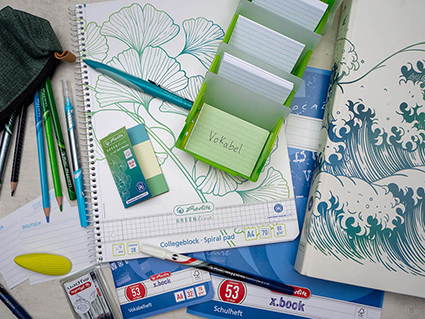 Verschieden Schulmaterialien der Marke Herlitz sind auf einem hellen Untergrund ausgebreitet: Ein karierter Collegeblock mit gezeichneten Ginko-Blättern, ein Ringbuch mit gezeichneten Wellen, ein Heftchen mit Haft-Memos, verschiedene Karteikarten, ein Mäppchen aus dem Stifte herausschauen, ein hellgrüner Radiergummi und ein Tintenlöscher. Foto: Johanna Rundel 
