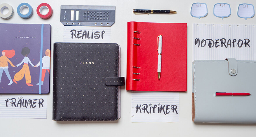 Auf einem weißen Untergrund liegen verschiedene Planer und Notizbücher der Marke Filofax. Sie sollen die Disney-Kreativitätstechnik symbolisieren. Von links nach rechts: Dunkelblaues Notizbuch mit diversen, bunten, tanzenden Frauen darauf, darunter der Zettel für die Rolle "Träumer", schwarzer Lederplaner mit goldenen Pünktchen, auf dem "Plans" steht, dazu ein Zettel mit der Aufschrift "Realist", ein knallrotes Clipbook, dazu ein Zettel mit dem Wort "Kritiker" und als letztes ein hellgrauer Planer mit dem dazugehörigen Zettel "Moderator". Um die Planer sind diverse, zur Rolle passende Gegenstände verteilt. Alles ist im Raster, sehr grafisch ausgelegt und von oben fotografiert. Foto: Johanna Rundel