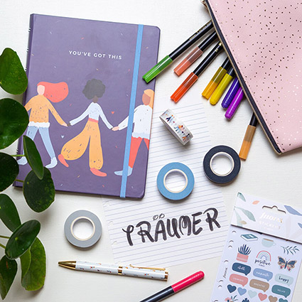 Auf einem weißen Untergrund liegt ein dunkelblaues Notizbuch mit diversen, bunten, tanzenden Frauen darauf, darunter ein ausgerissener, linierter Zettel mit dem schnörkeligen Wort "Träumer". Drumherum sind verschiedene Materialien verteilt: ein rosa, ein klippbares Mäppchen von Filofax, bunte Fasermaler, Washitapes, Aufkleber ein Kugelschreiber. Von links ragt eine kleine Zimmerpflanze ins Bild. Foto: Johanna Rundel