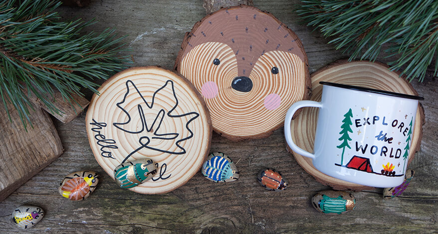 Drei Baumscheiben liegen auf einem rustikalen Holzuntergrund. Auf der ersten Scheibe ist mit schwarzem Stift von edding die Outline eines Ahornblattes gemalt. Auf der mittleren ist das Gesicht eines Fuchses zu sehen. Auf der rechten Baumscheibe liegt eine bemalte Emaille-Tasse mit Camping-Motiv. Im unteren Drittel des Bildes liegen mehrere Steine in einer waagrechten Reihe. Sie wurden als Käfer bemalt. Rund um die Objekte sind Holzscheite und Kiefernzweige zu sehen. Foto: Johanna Rundel
