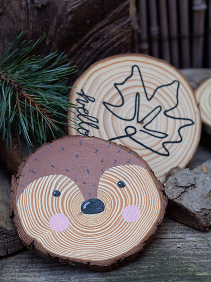 Zwei Baumscheiben liegen auf einem rustikalen Holzuntergrund. Auf der vorderen Scheibe ist das Gesicht eines Fuchses zu sehen. Auf der mittleren Baumscheibe ist mit Hilfe eines schwarzen Stifts von edding die Outline eines Ahornblattes gemalt. Rund um die Scheiben sind Holzscheite und Kiefernzweige zu sehen. Foto: Johanna Rundel