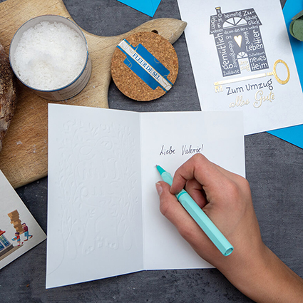 Jemand schriebt mit einem türkisfarbenen Tintenroller in eine Grußkarte des ABC Glückwunschkartenverlags. Eine weitere Karte zum Umzug liegt in der rechten oberen Ecke. Oberhalb der Karte ist ein Holzbrett zu sehen, auf dem Brot und Salz stehen. Foto: Johanna Rundel