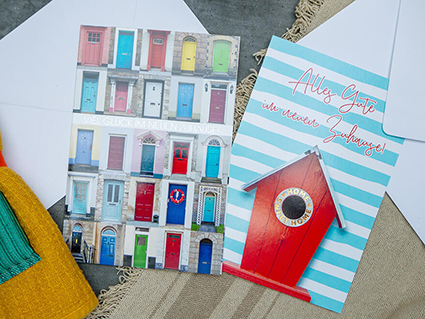 Auf einem anthrazitfarbenen Untergrund liegen zwei Grußkarten des ABC Glückwunschkartenverlag. Auf der linken Karte sind Fotos von sehr vielen bunten Hauseingangstüren zu sehen. Dazwischen steht: "Viel Glück im neuen Zuhause". Auf der rechten Karte, mit türkis-weißen Querstreifen, ist ein rotes Vogelhaus abgebildet. Der Schriftzug darüber lautet: "Alles Gute im neuen Zuhause!". Foto: Johanna Rundel