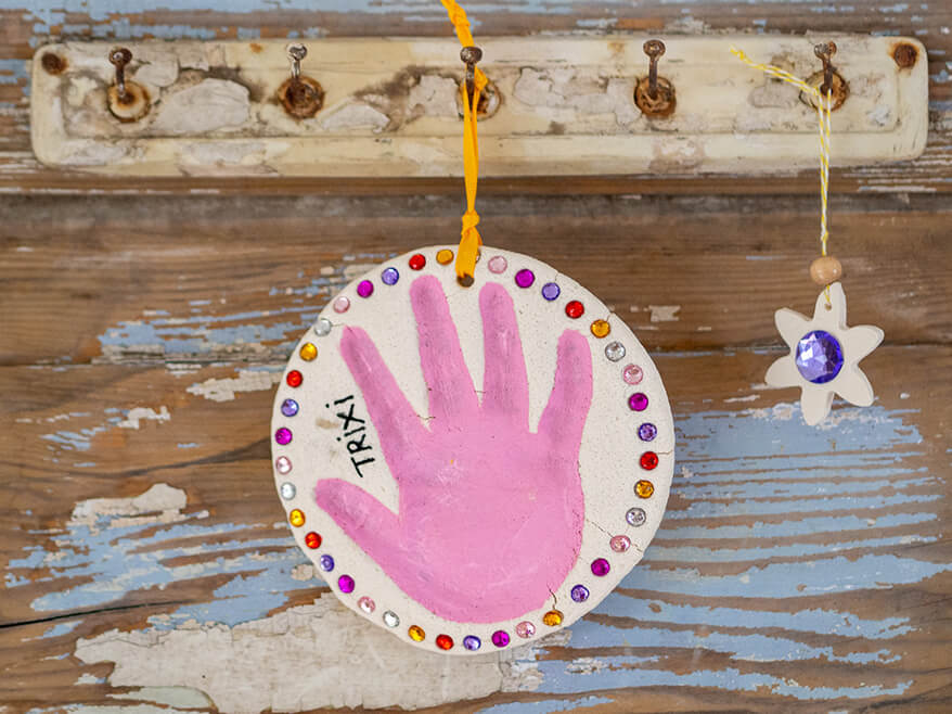 An einer Hakenleiste an einer Holztür – beides im Shabby-Chic – hängen zwei Objekte aus Salzteig. Eine runde Scheibe mit Handabdruck und Glitzersteinen von Knorr Prandell, sowie ein kleiner Blumenanhänger mit blauem Glitzerstein in der Mitte. Foto: Johanna Rundel 