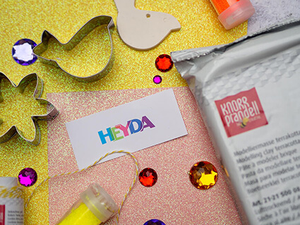 Auf dem Bild sind verschiedene Bastelmaterialien der Marken Heyda und Knorr Prandell zu sehen: Lufttrocknende Modelliermasse, Glitzersteine, Glitter, Ausstechförmchen und Glitzerkarton in Gelb und Orange. Foto: Johanna Rundel 