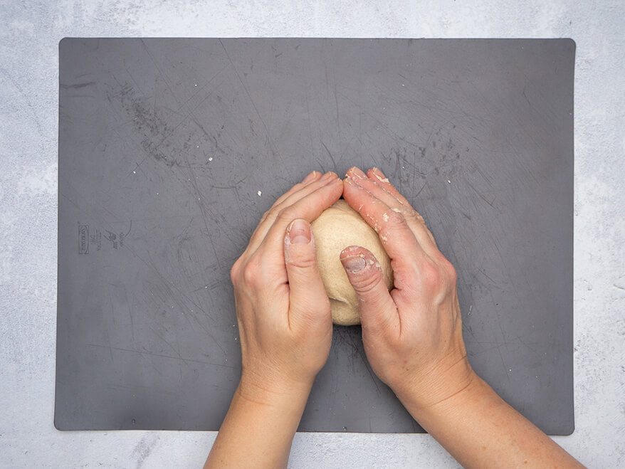 Jemand rollt auf einer dunkelgrauen Backmatte zwischen den Händen einen Ball aus einer Portion Salzteig. Der Untergrund ist Betongrau. Foto: Johanna Rundel 