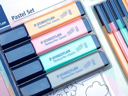 Das Bild zeigt einen Ausschnitt des Pastel Set von Staedtler in Originalverpackung. Zu sehen sind vier breite Textmarker in Orange, Rosa, Türkis und Grau, sowie vier dünne Textmarkerstifte in denselben Farben. Außerdem sieht man einen Aufkleberbogen mit Stickern zum Ausmalen. Foto: Johanna Rundel