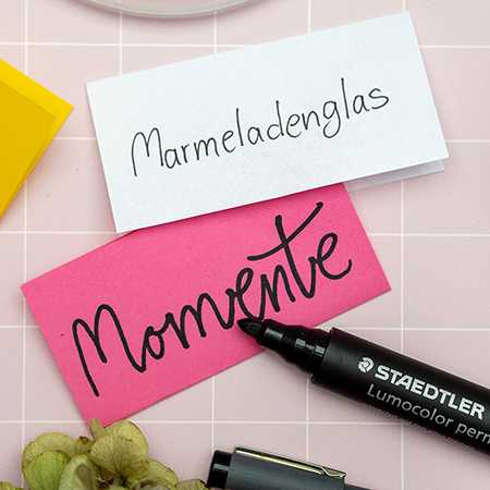 Auf einem rosafarbenen Untergrund mit weißem Karomuster liegen ein weißer und darunter ein pinker Notizzettel. Auf dem Weißen steht: „Marmeladenglas“, auf dem Pinken: „Momente“. Auf dem pinken Zettel liegt ein geöffneter, schwarzer Permanentmarker von Staedtler. Foto: Johanna Rundel