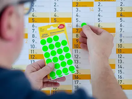 Ein dunkelhaariger Mann mit Brille klebt neongrüne Klebepunkte von Avery Zweckform auf einen großen Wandkalender. Foto: Johanna Rundel