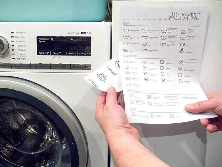 Jemand befestigt einen Hinweis zu Waschsymbolen auf einem wiederablösbaren Etikett von Avery Zweckform neben einer Waschmaschine. Foto: Johanna Rundel