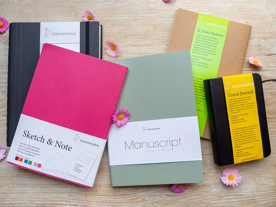 Auf einem rustikalen Holzbrett liegen verschiedene Skizzen- und Notizbücher, Travel-Journals und Booklets von Hahnemühle.