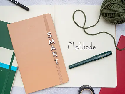 Ein rotes, ein rosa und ein waldgrünes Notizbuch von Moleskine liegen nebeneinander. Am Gummiband des rosa Buches wurde die silbernen Buchstaben S, M, A, R und T befestigt. Foto: Johanna Rundel