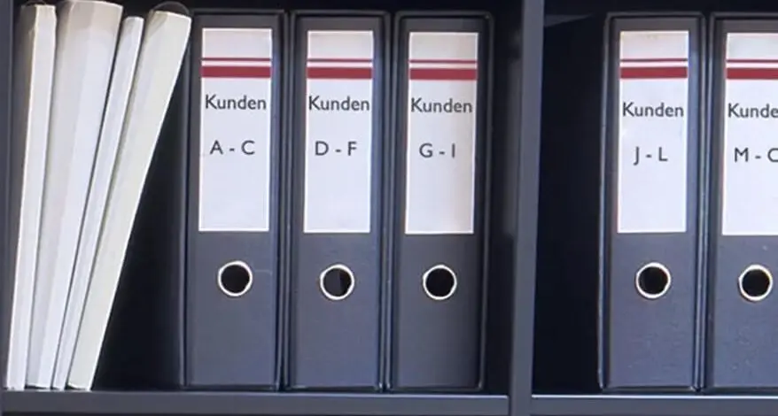 Viele schwarze Aktenordner stehen in alphabetischer Reihenfolge in einem schwarzen Aktenregal.