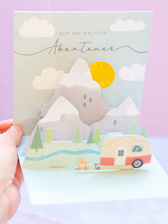 Jemand hält eine Pop-Up-Karte von Grafik Werkstatt mit einem Bergmotiv, vor dem ein Wohnwagen steht und dem passenden Spruch „Auf ins nächste Abenteuer“ in die Kamera. Foto: Johanna Rundel