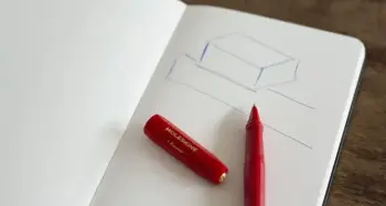 Ein roter Kugelschreiber liegt auf einem aufgeschlagenen Notizbuch, in das einfache Quader gezeichnet sind. 