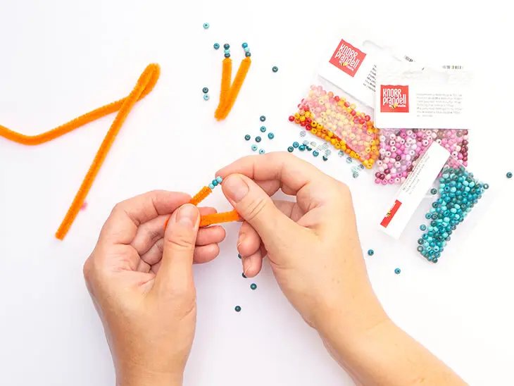 Jemand fädelt kleine blaue Holzperlen auf die Enden von neonorange-farbigen Pfeifenputzern von KNORR prandell. Foto: Johanna Rundel