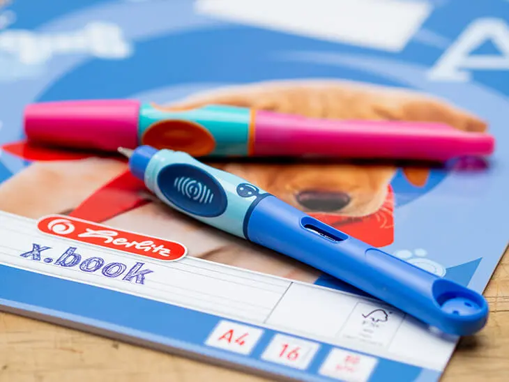 Ein rosa und ein blauer Schreiblernfüller von Pelikan liegen auf einem DIN-A4-Heft für die dritte Klasse von herlitz. Foto: Johanna Rundel