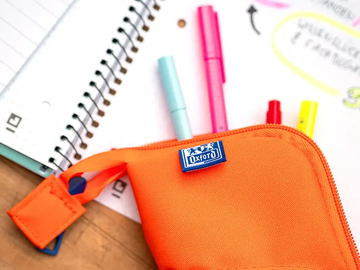 Ein orangefarbenes Mäppchen von Oxford liegt auf einem aufgeschlagenen Collegeblock, mehrere Stifte schauen hervor. Foto: Johanna Rundel