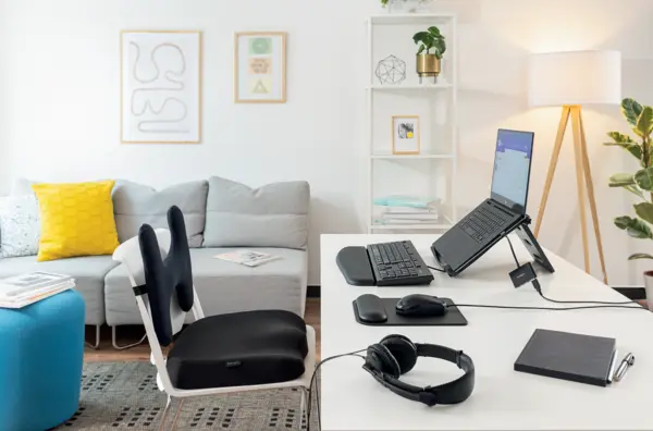 Das Bild zeigt einen ergonomisch gestalteten Arbeitsplatz mit einem Laptop auf einem Ständer, einer externen Tastatur, einer Maus und ergonomischen Mauspads sowie Handgelenkauflagen von Kensington. Ergänzt wird die Einrichtung durch ein komfortables Sitzkissen und eine Rückenstütze auf einem Stuhl.