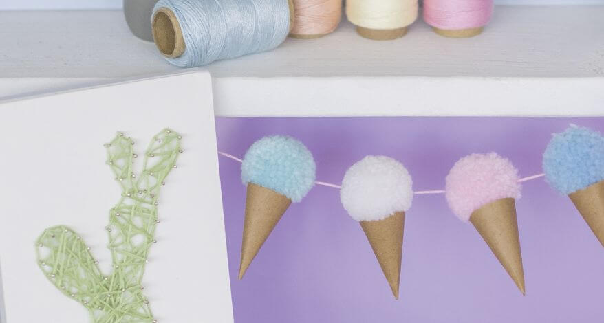 Nahaufnahme einer Rageldekoration: Auf einer Kordel sind selbstgebastelte Eistüten aus Kraftpapier und Pompons zu sehen, gebastelt mit Materialien der Marke Knorr Prandell. Daneben sieht man den Ausschnit eines Kaktusses, der als Fadenbild gestaltet wurde. Auf einem Regalbrett darüber sieht man verschiedene Garnrollen in Pastellfarben.