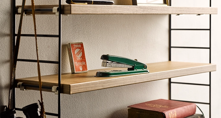 Das Bild zeigt ein schlichtes Holzregal. Auf einem der Regalböden steht ein grünes, vintage Heftgerät und eine kleine Schachtel mit der Aufschrift „Novette“, darunter liegt ein dickes, altes Buch. Die Szene erinnert an ein Arbeitszimmer aus vergangenen Zeiten.