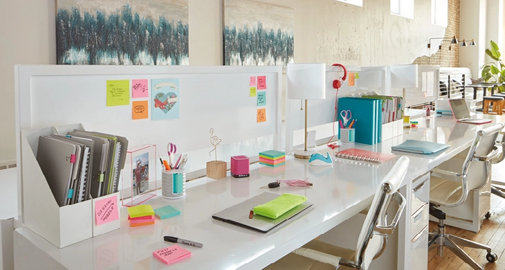 In einem hellen Büro steht ein weißer Schreibtisch mit Post-it®-Blöcken in verschiedenen Farben und Formaten verteilt. An der Rückwand hängen beschriftete Post-it®-Haftnotizen. In einem Stehsammler befinden sich mit Post-it® Page Markern gekennzeichnete Blöcke. Links oben in der Grafik ist das Post-it® Logo integriert.