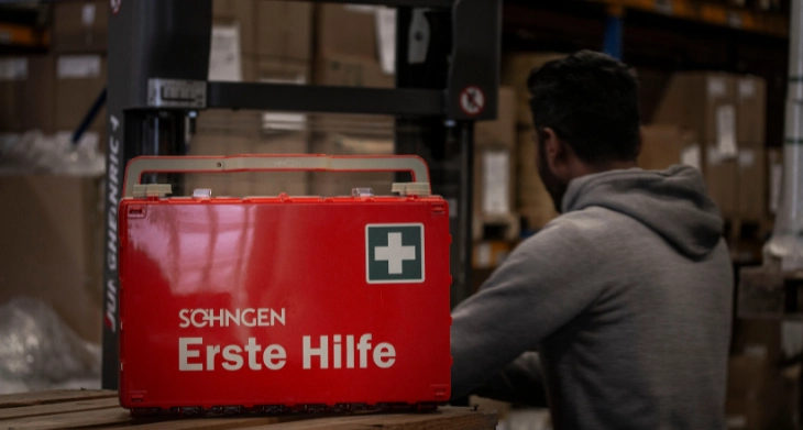 Roter Erste-Hilfe-Kasten mit weißem Kreuz in einem Lager, Mann im Hintergrund. 