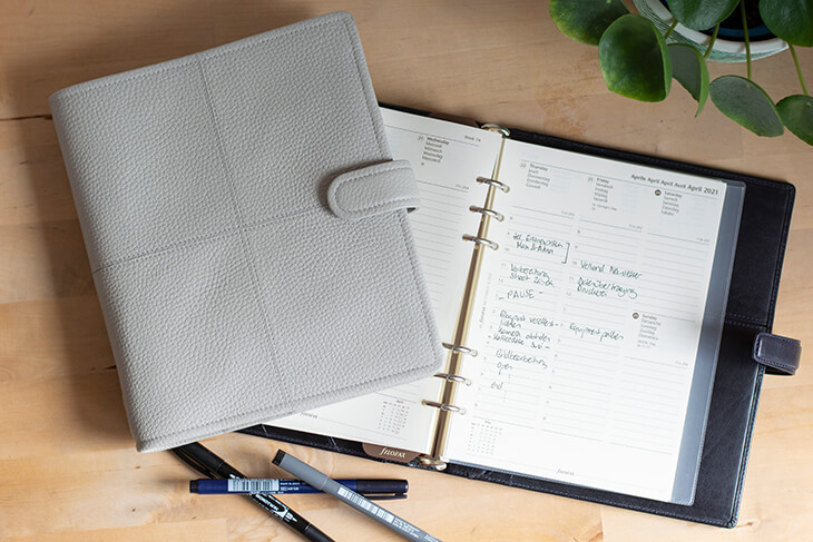 Aus der Vogelperspektive blickt man auf zwei Kalender von Filofax. Einer mit schwarzem Einband liegt aufgeklappt auf dem Schreibtisch, einer mit hellgrauem Ledereinband liegt zusammengeklappt schräg darauf. Man sieht einige Einträge auf einer Seite und Fineliner am unteren Bildrand.