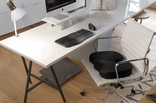 Das Bild zeigt einen modernen Arbeitsplatz mit einem Monitorständer, der den Bildschirm auf Augenhöhe positioniert. Ergänzt wird der ergonomische Arbeitsplatz durch eine externe Tastatur, Maus, ein Sitzkissen auf dem Bürostuhl und eine Fußstütze.