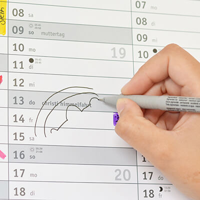 Detailaufnahme eines Wandkalenders: Eine Hand zeichnet mit einem Fineliner eine Brücke über den Brückentag an Christi Himmelfahrt.