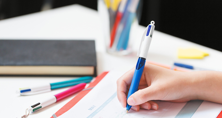 Hand schreibt mit blauem BIC 4 Colours Kugelschreiber in einen Kalender, umgeben von weiteren BIC-Stiften und einem Notizbuch auf einem Schreibtisch.