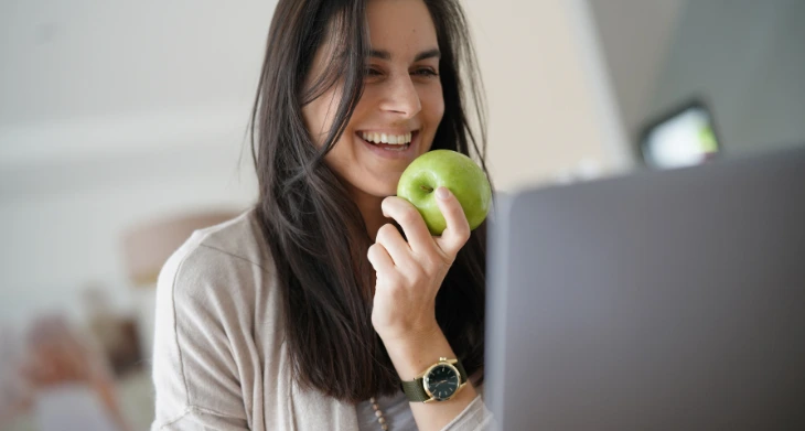 Lachende Frau sitzt mit Apfel in der Hand vor einem Laptop