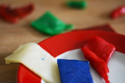 Mehrere Streifen Knetbienenwachs in den Farben rot, blau, weiß und grün leigen auf einem Teller und dem Tisch.