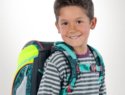 Ein Junge steht vor einer weißen Wand und lächelt in die Kamera. Am Rücken trägt er einen Ranzen der Marke Scout.