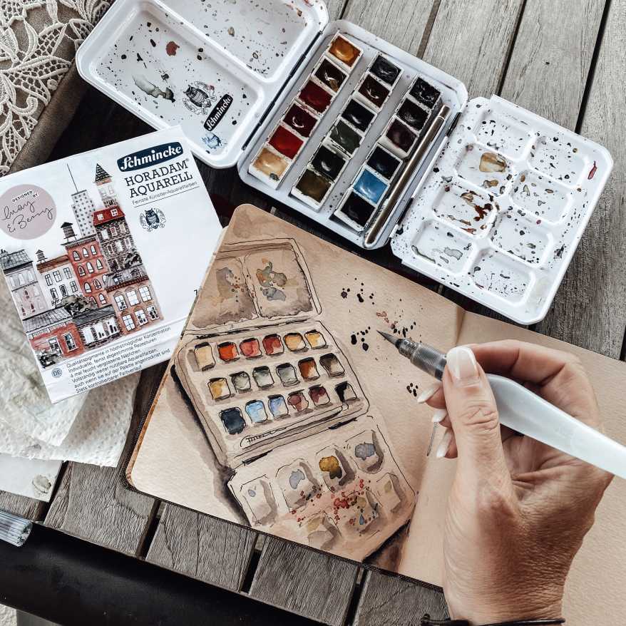 Ein braun getöntes Skizzenbuch der Makre Hahnemühle mit einem aqaurellierten Aquarellfarbkasten. Neben dem Buch liegt ein Aquarellkasten der Marke Schmicke.