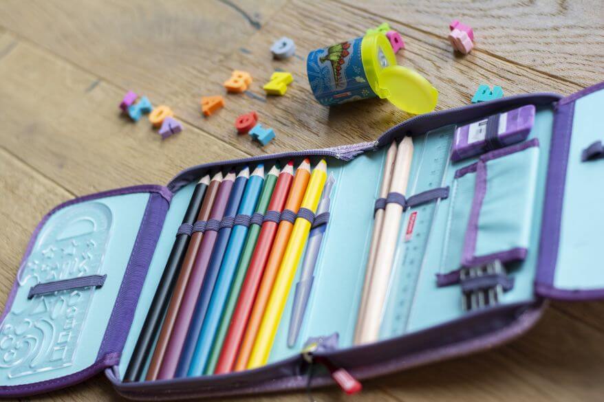 Ein geöffnetes Schulmäppchen mit Buntstiften liegt auf einem Schreibtisch. Dahinter liegt ein Anspitzer mit Radiergummis in ABC-Buchstabenform.
