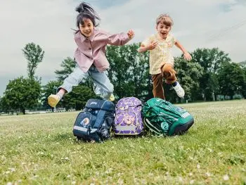 Zwei Kinder hüpfen auf einer grünen Wiese über ihre Schulranzen von ergobag.