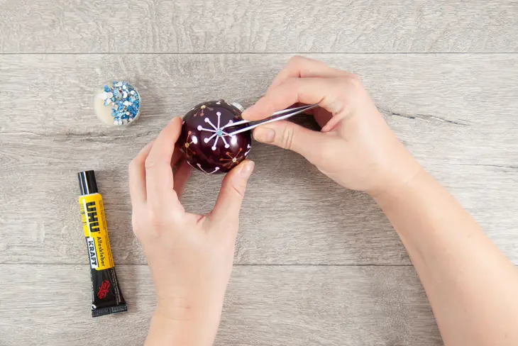 Eine Hand setzte mit einer Pinzette Glitzersteine auf eine selbst gestaltete Weihnachtskugel