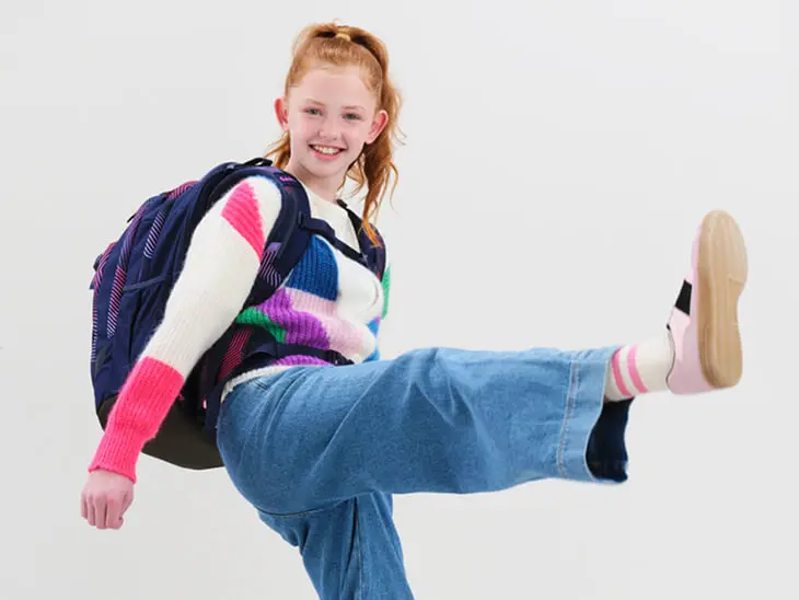 Eine Jugendliche mit einem Schulrucksack von satch kickt fröhlich ihr Bein nach oben.
