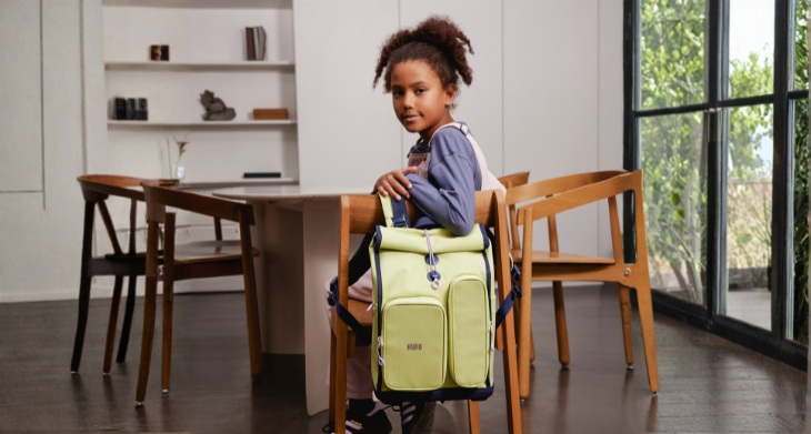 Ein Mädchen sitzt an einem Tisch und dreht sich zur Kamera, über ihrem Stuhl hängt ein Schulranzen von KWIO