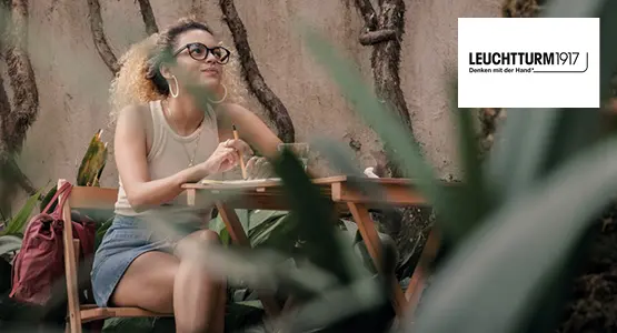 Eine junge Frau, die mit einem Notizbuch von LEUCHTTURM1917 an einem Holztisch sitzt und nachdenkt.