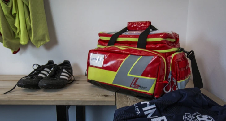 Erste-Hilfe-Tasche Lifebag auf Holzbank in Umkleide mit Fußballschuhen und Trikot.